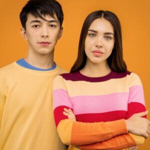 A young couple standing and posing together in casual colorful sweaters against an orange background.