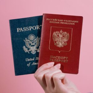 A hand holding US and Russian passports against a pink background.