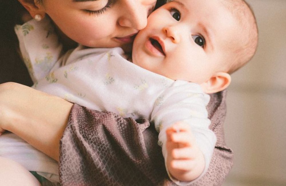 A mother lovingly embraces her smiling baby indoors in Minsk, Belarus.